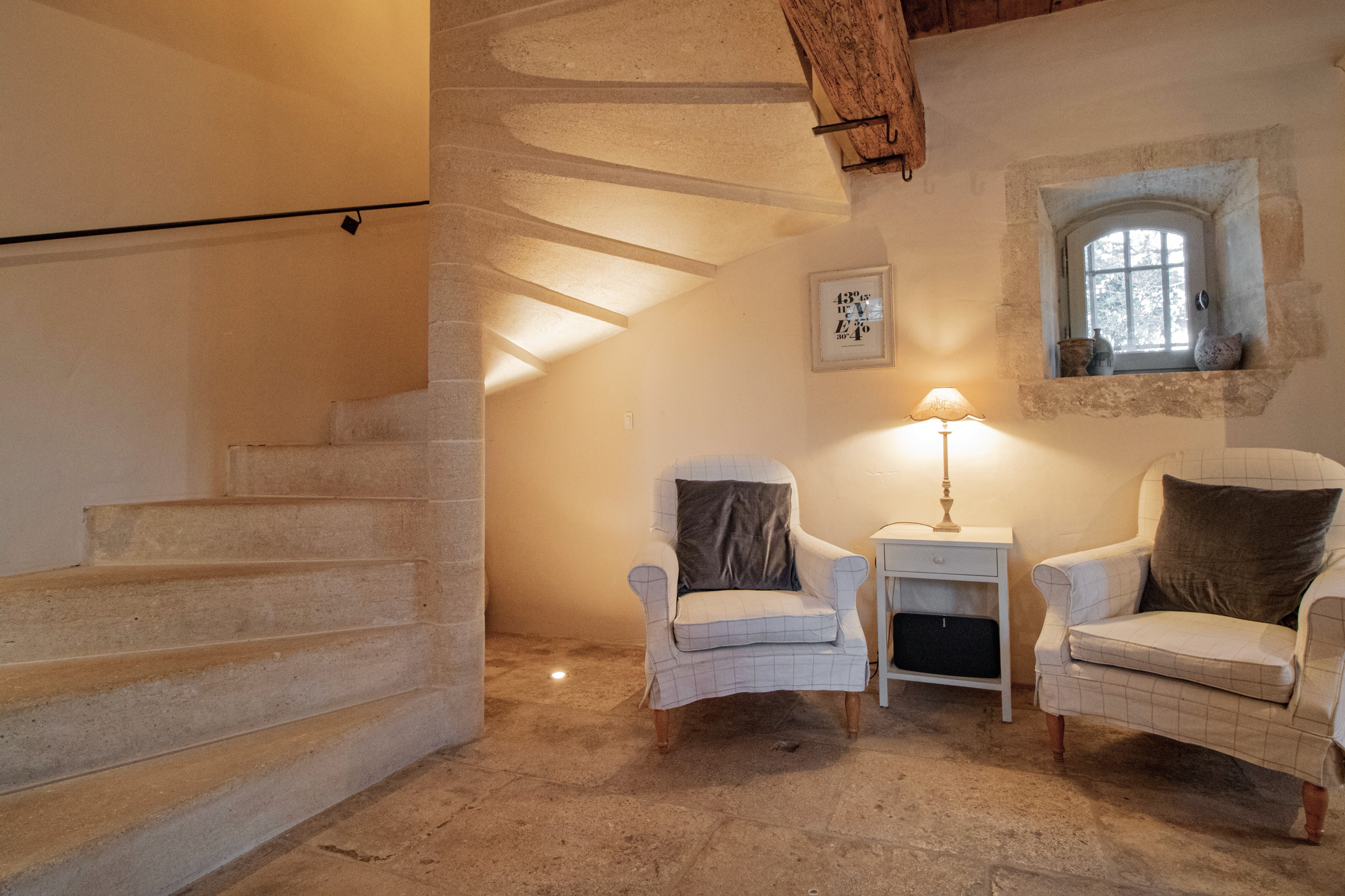 An interior room featuring a stone staircase to the left and two armchairs with striped upholstery near a small window. There is a lamp on a white side table between the chairs, with beige walls and a wooden beam overhead.