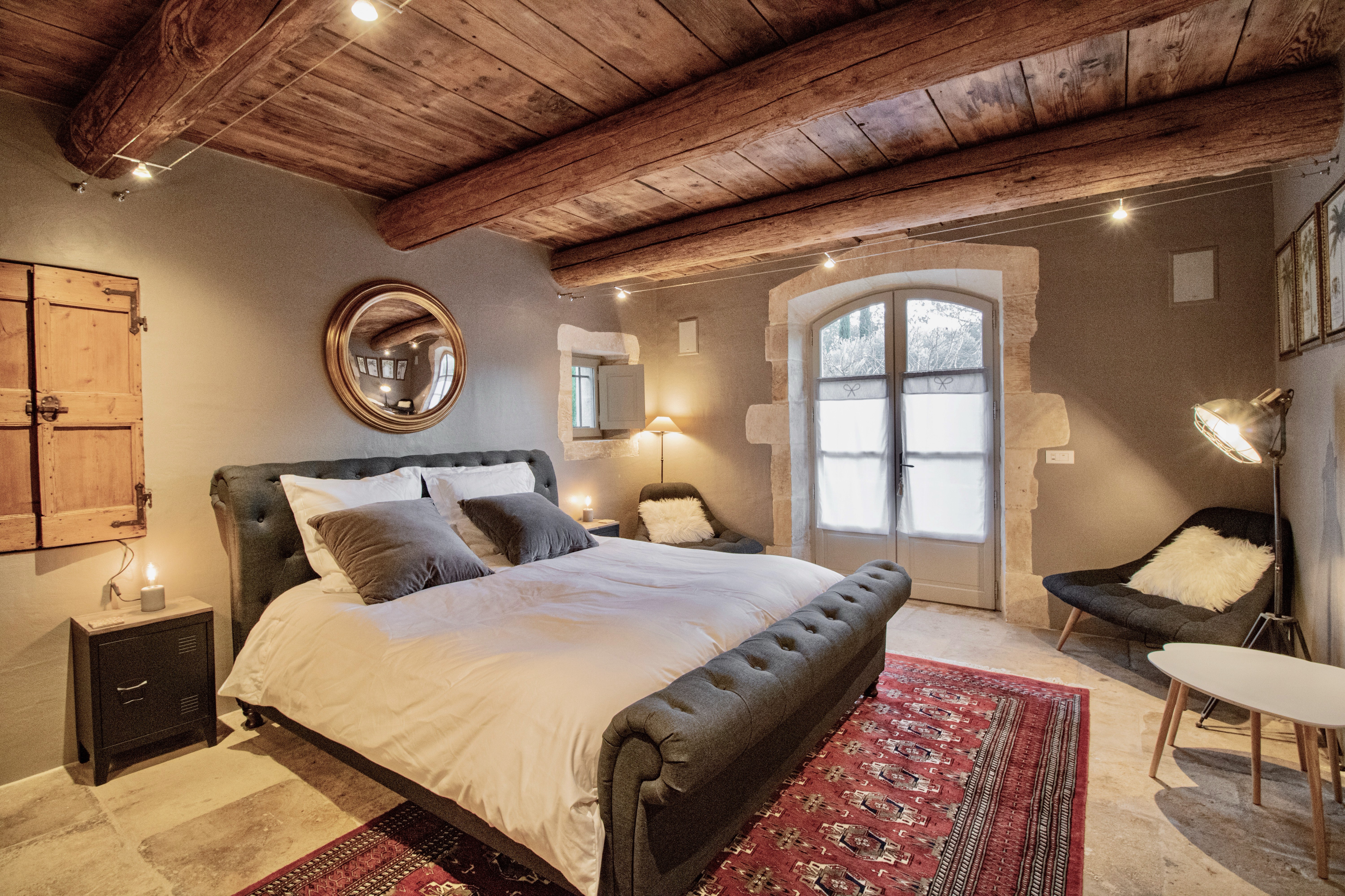 A cozy, rustic bedroom featuring a large bed with gray bedding, exposed wooden beams, a red patterned rug, and a round mirror on the wall. Natural light filters through arched glass doors.