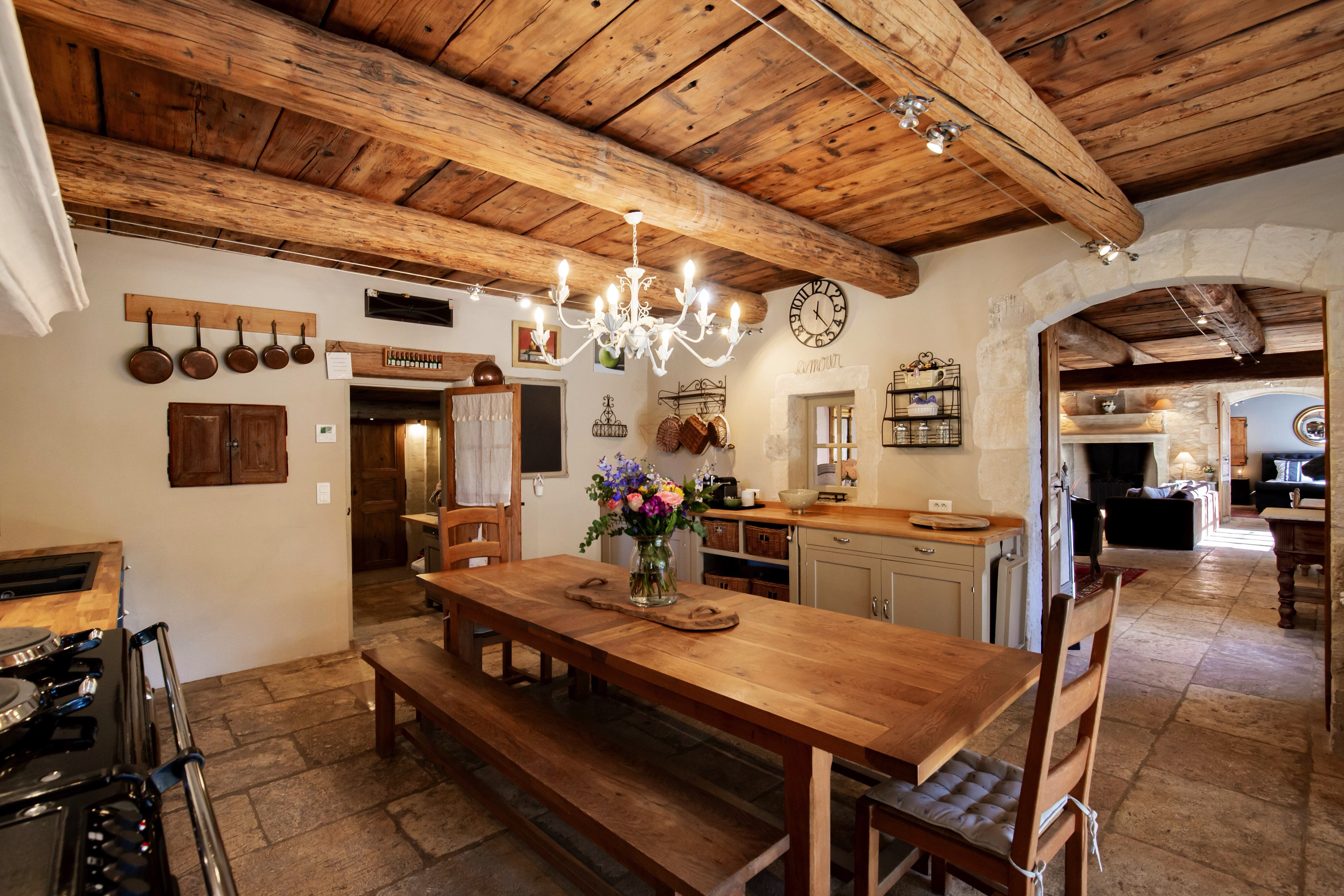 A rustic kitchen with exposed wooden beams, a large dining table with benches, vintage decor, and a chandelier. a cozy living area is visible through an archway.