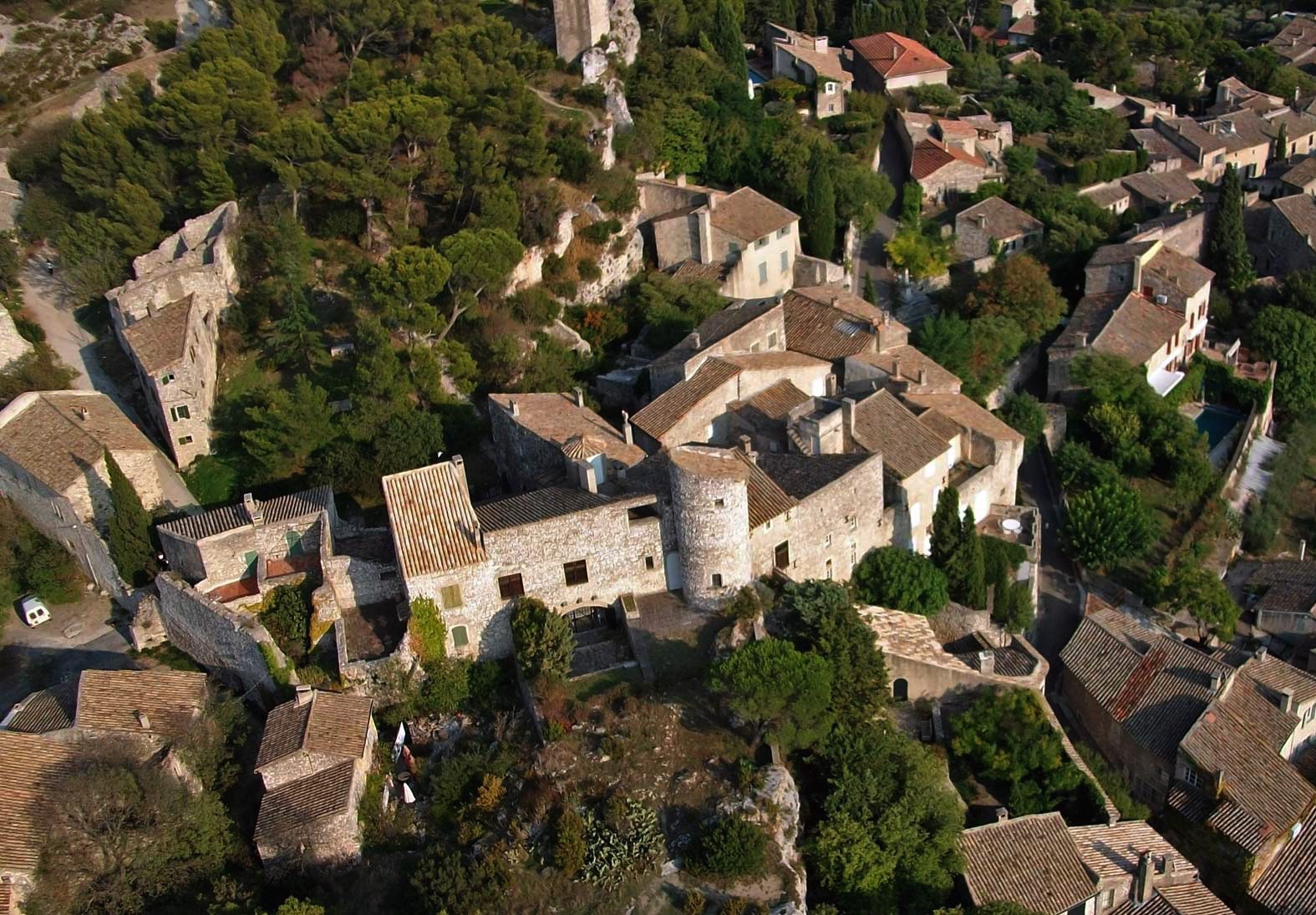 Aerial view of a historic village with tightly clustered stone buildings surrounded by lush greenery and trees under a clear sky.