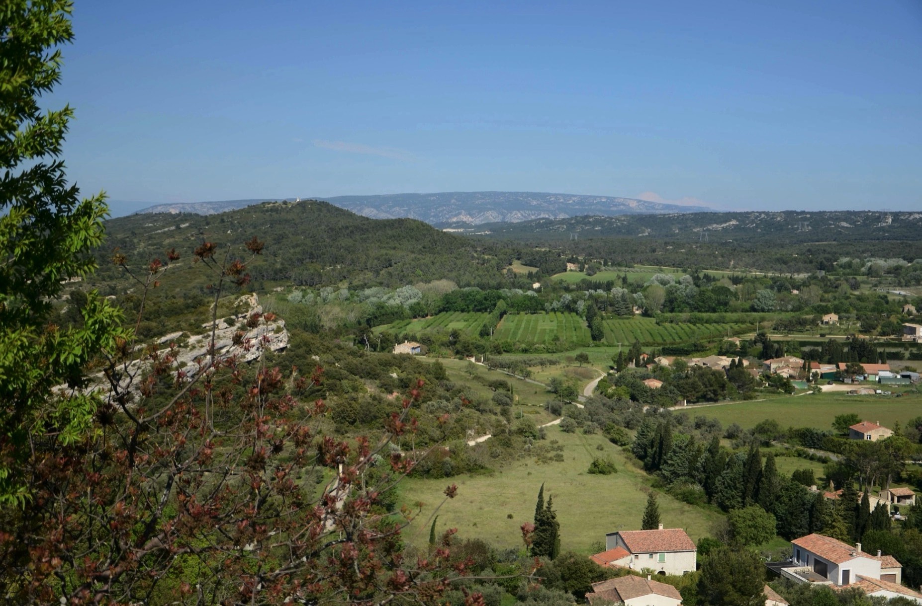 Panoramic view of a lush, hilly landscape with scattered houses, vineyards, and a winding road, under a clear blue sky.