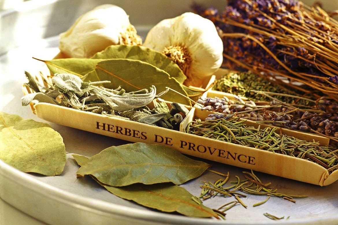 Assortment of dried herbs labeled 'herbes de provence', including rosemary, thyme, bay leaves, and lavender, alongside garlic bulbs on a sunlit table.