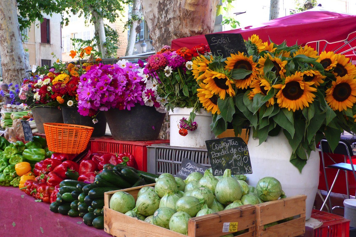 A vibrant outdoor market display featuring an array of fresh vegetables, sunflowers, and assorted colorful flowers under a sunny backdrop.